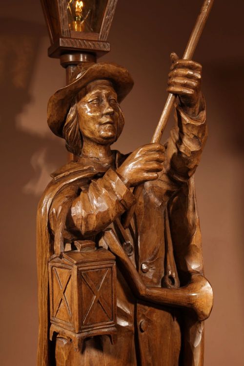 fig 1. A hand-carved wooden sculpture of a night watchman holding a horn and halberd beside an illuminated street lantern on a stacked timber base.