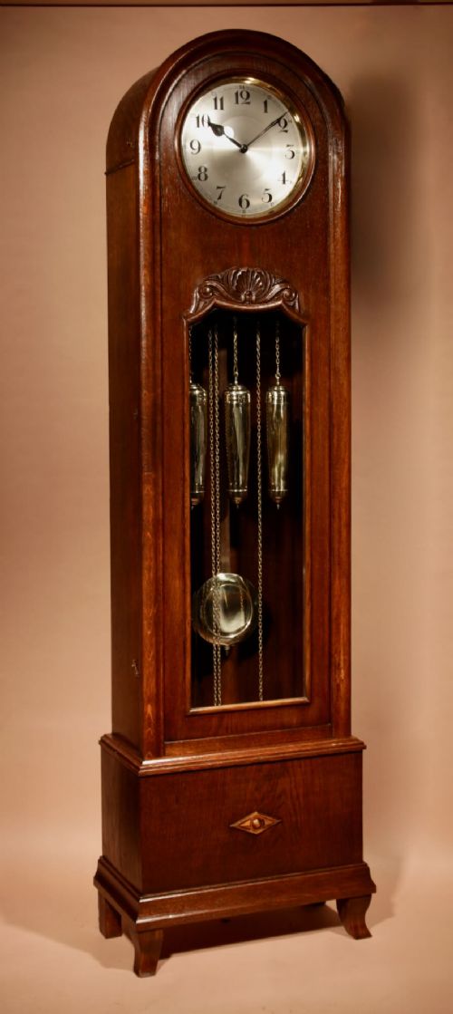 fig 1. An Art Deco dark oak longcase clock featuring an arched hood, silvered dial with Arabic numerals, glazed trunk door showcasing weights, and a carved geometric base.