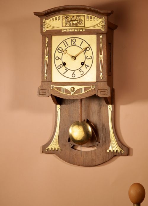 fig 1. An Art Nouveau light oak wall clock featuring brass mounts, an embossed chariot motif top panel, and a visible pendulum.