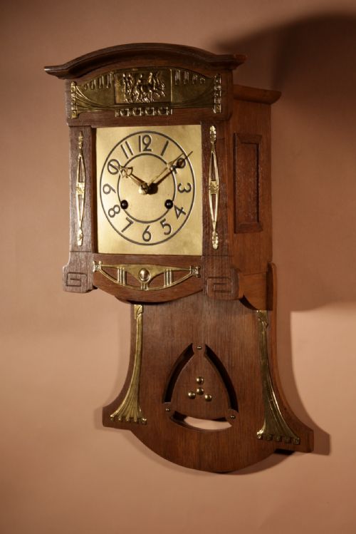 fig 2. An Art Nouveau light oak wall clock featuring brass mounts, an embossed chariot motif top panel, and a visible pendulum.