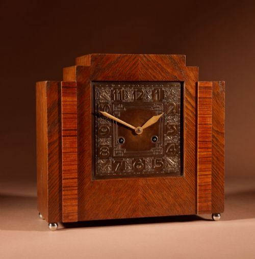 fig 1. An Art Deco mantel clock crafted from oak and rosewood veneer featuring a stepped architectural top, a square brass dial with relief numerals, and small metal ball feet.