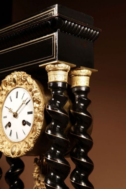 fig 1. An ebonised French portico clock featuring brass stringing inlay, four barley-twist columns, an ornate gilt dial bezel, and an exposed ornate pendulum.