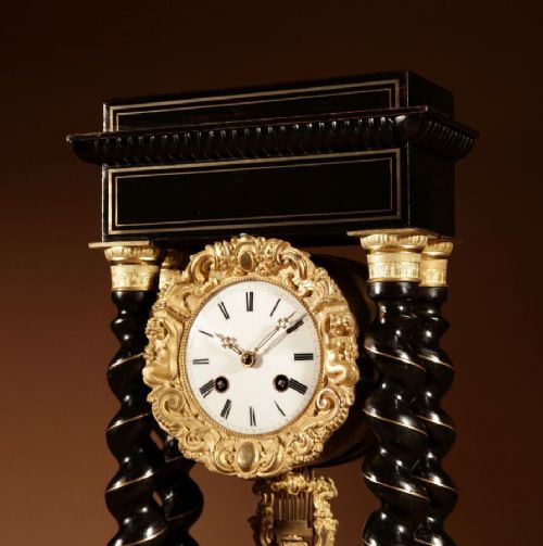 An ebonised French portico clock featuring brass stringing inlay, four barley-twist columns, an ornate gilt dial bezel, and an exposed ornate pendulum. View V