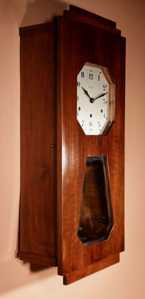 An Art Deco French walnut wall clock featuring a stepped top, an octagonal white dial face with Arabic numerals, and a visible brass pendulum bob behind a trapezoidal glass aperture. View IX