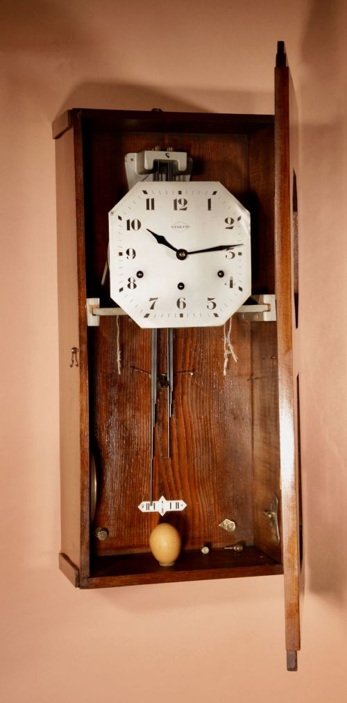 An Art Deco French walnut wall clock featuring a stepped top, an octagonal white dial face with Arabic numerals, and a visible brass pendulum bob behind a trapezoidal glass aperture. View X