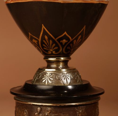 A 19th-century Grand Tour table lamp featuring a black ceramic urn base with red-figure chariot artwork, gilt bronze handles, and a pleated conical fabric shade. View X