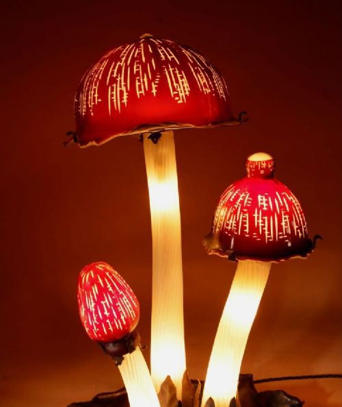 fig 1. A triple mushroom table lamp featuring three illuminated white glass stems topped with red patterned glass caps set into a dark wrought iron base.
