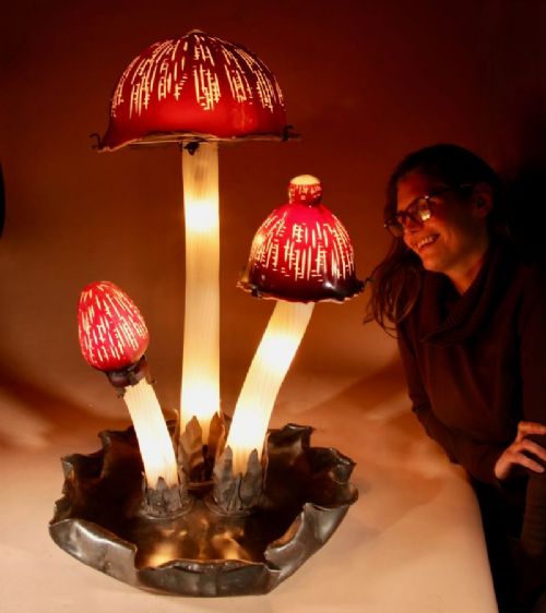 A triple mushroom table lamp featuring three illuminated white glass stems topped with red patterned glass caps set into a dark wrought iron base. View VII
