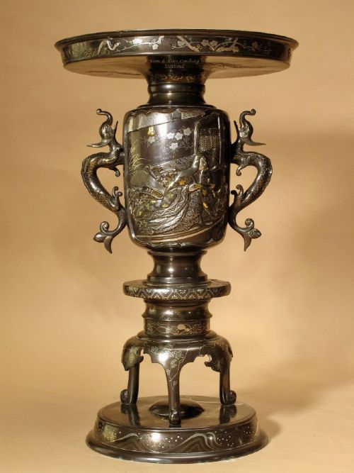 fig 1. A pair of dark Japanese bronze vases featuring relief figures, gold and silver inlay, dragon-shaped side handles, and broad flat rims on integrated footed bases.