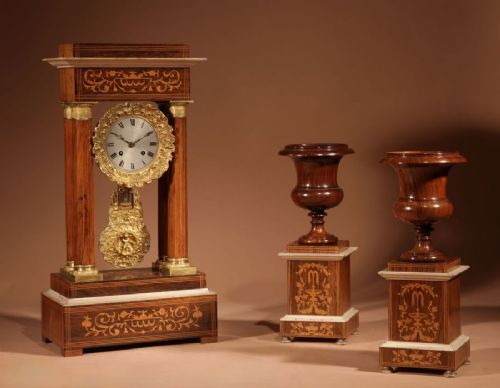 fig 1. A French Louis Philippe portico mantel clock set featuring floral wood inlay, architectural columns, a gilt metal pendulum, and two matching urn side pieces on decorative pedestals.