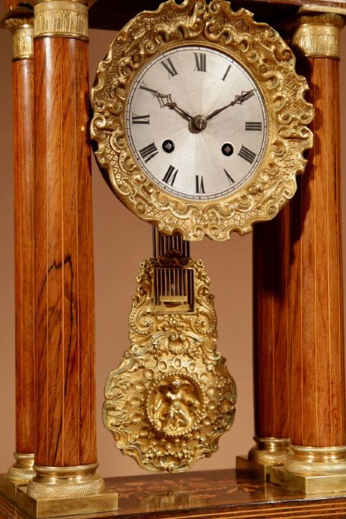 fig 3. A French Louis Philippe portico mantel clock set featuring floral wood inlay, architectural columns, a gilt metal pendulum, and two matching urn side pieces on decorative pedestals.
