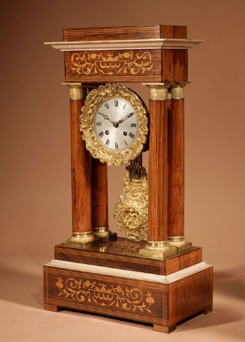 A French Louis Philippe portico mantel clock set featuring floral wood inlay, architectural columns, a gilt metal pendulum, and two matching urn side pieces on decorative pedestals. View XV
