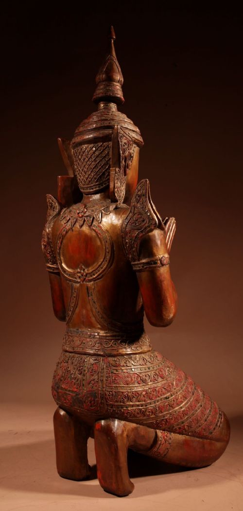 A carved wooden Thai Thepphanom guardian statue depicted kneeling with hands held in a prayer gesture, featuring a conical headdress and gilded red lacquered details. View XII