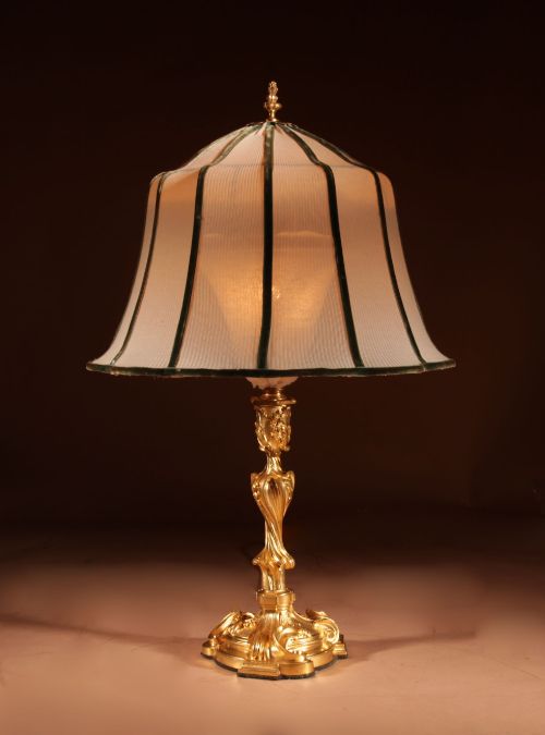 fig 1. A French Rococo style gilded bronze table lamp featuring an asymmetrical scrolling base and a fitted fabric shade with dark green vertical trim.