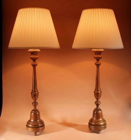 fig 1. A pair of tall patinated brass candlestick table lamps featuring turned stems and pleated cream shades on circular bases.