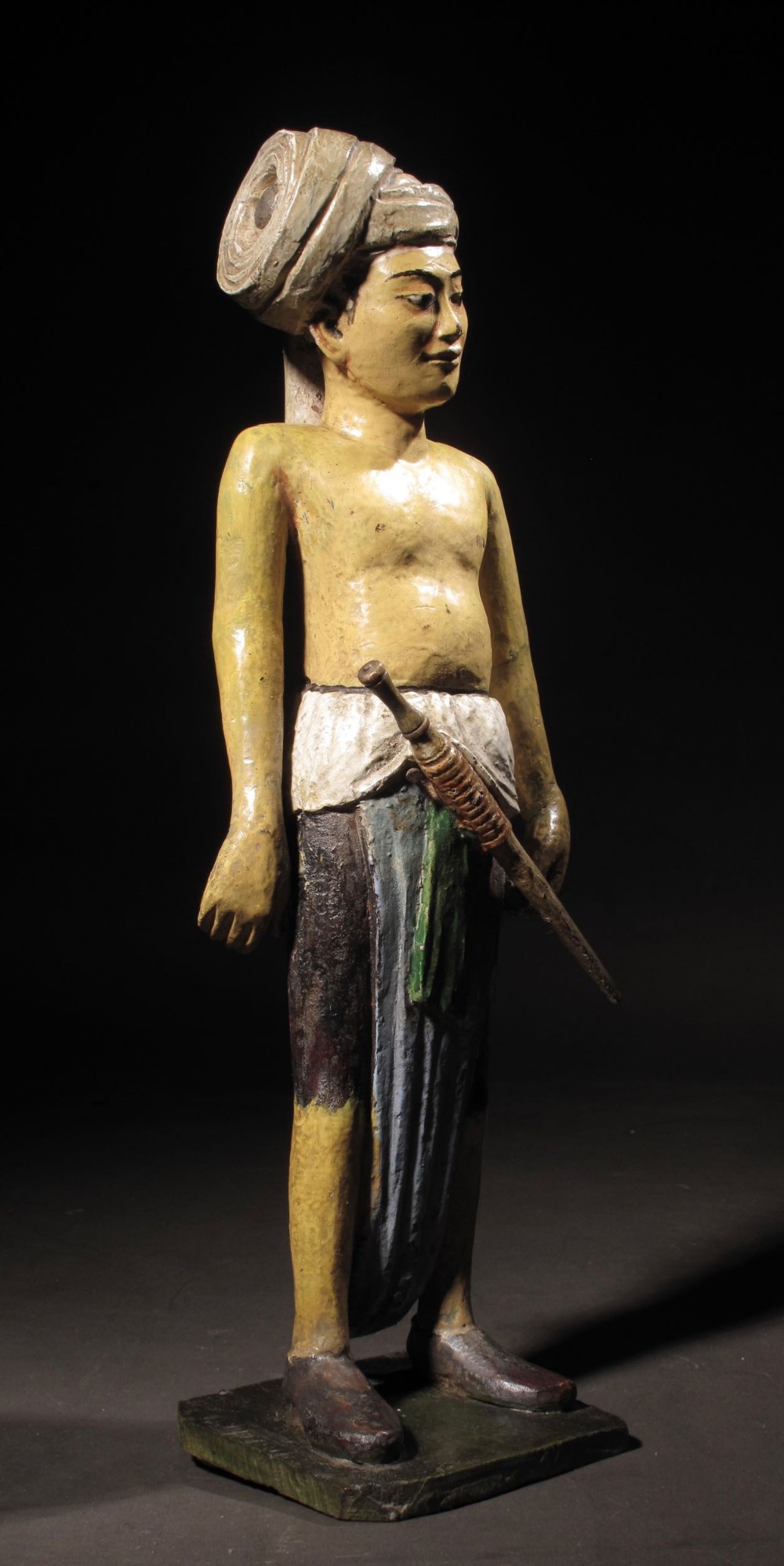 A carved wooden figure depicting a standing man with a large turban, bare torso, wrapped dhoti, and a weapon at his waist, painted in yellow, grey, and green pigments on a square base.