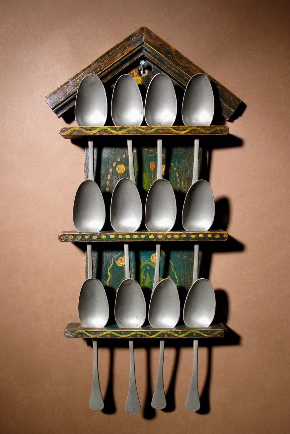 A dark green painted wooden spoon rack featuring three tiered shelves holding twelve pewter spoons, accented with hand-painted yellow floral motifs and a gabled top.