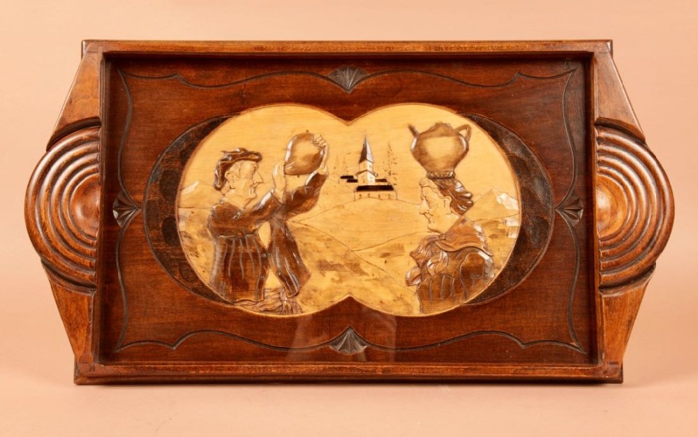 A rectangular warm brown carved wooden tray featuring stylized Art Deco ribbed handles and a central inlaid relief scene of two figures and a village church.