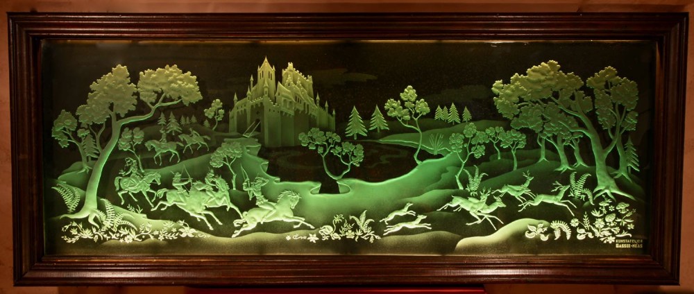 A large rectangular dark wood frame housing an illuminated green sandblasted glass panel featuring a detailed hunting scene with horseback archers, hounds, stags, and a castle.