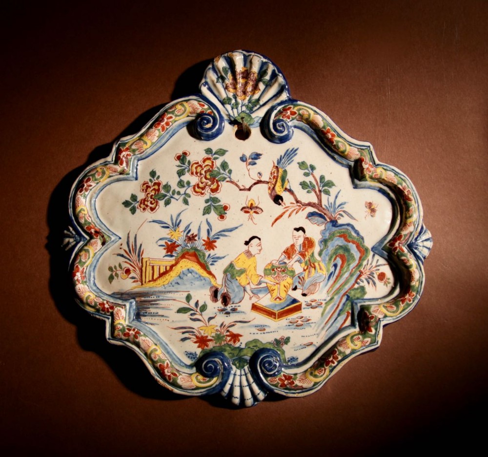 An 18th-century Dutch Delft tin-glazed earthenware plaque with a moulded shell border, decorated in polychrome enamel with figures in a Chinoiserie landscape.