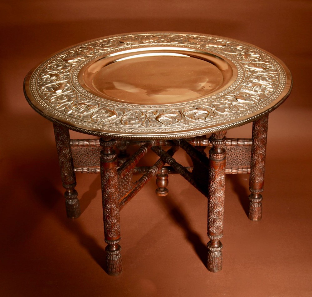 A circular hand-chased brass tray top featuring embossed foliate motifs rests on an intricate carved dark hardwood folding base.