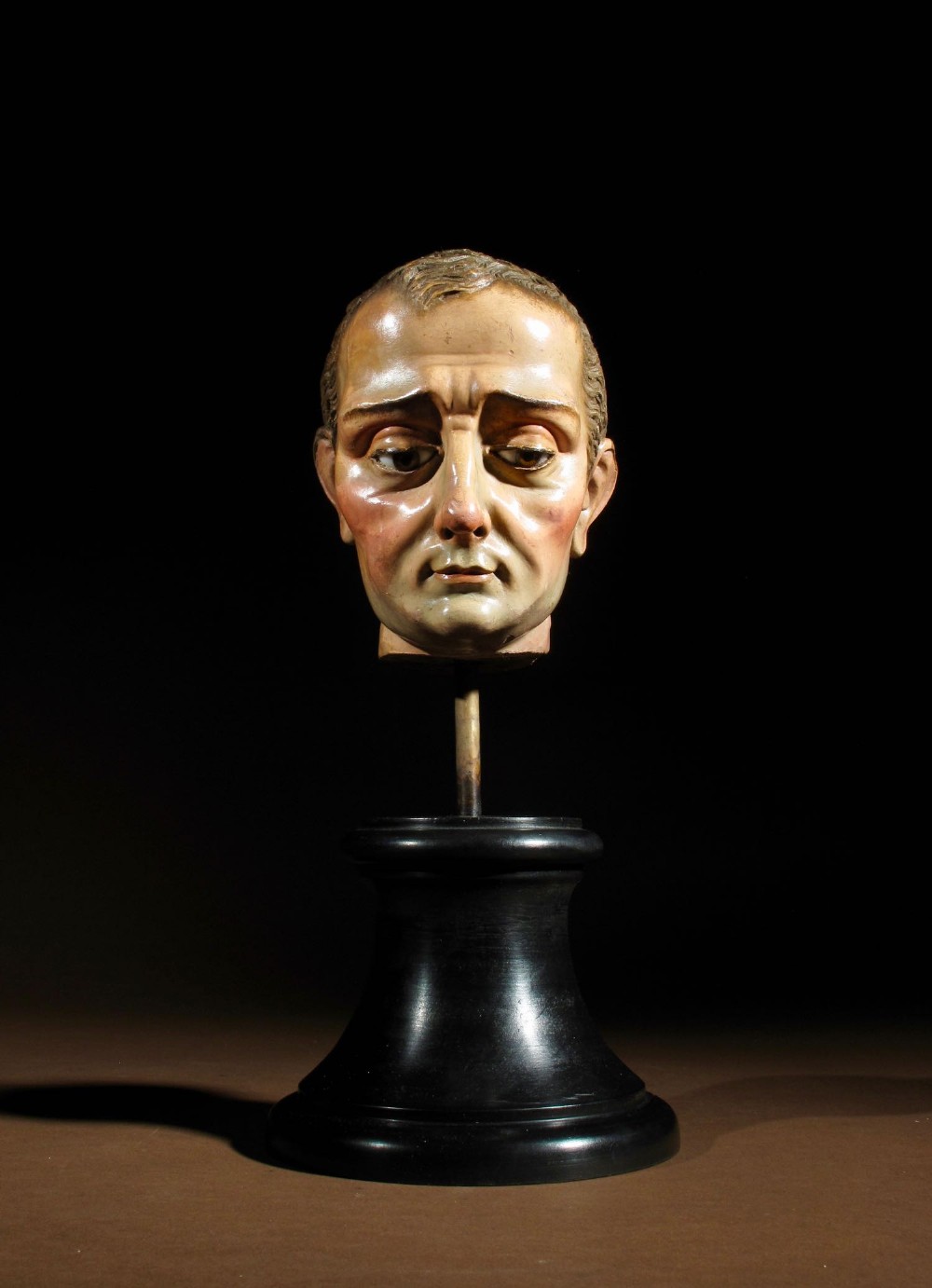 A carved and hand-painted polychrome wood head of a saint mounted on a black circular pedestal base.