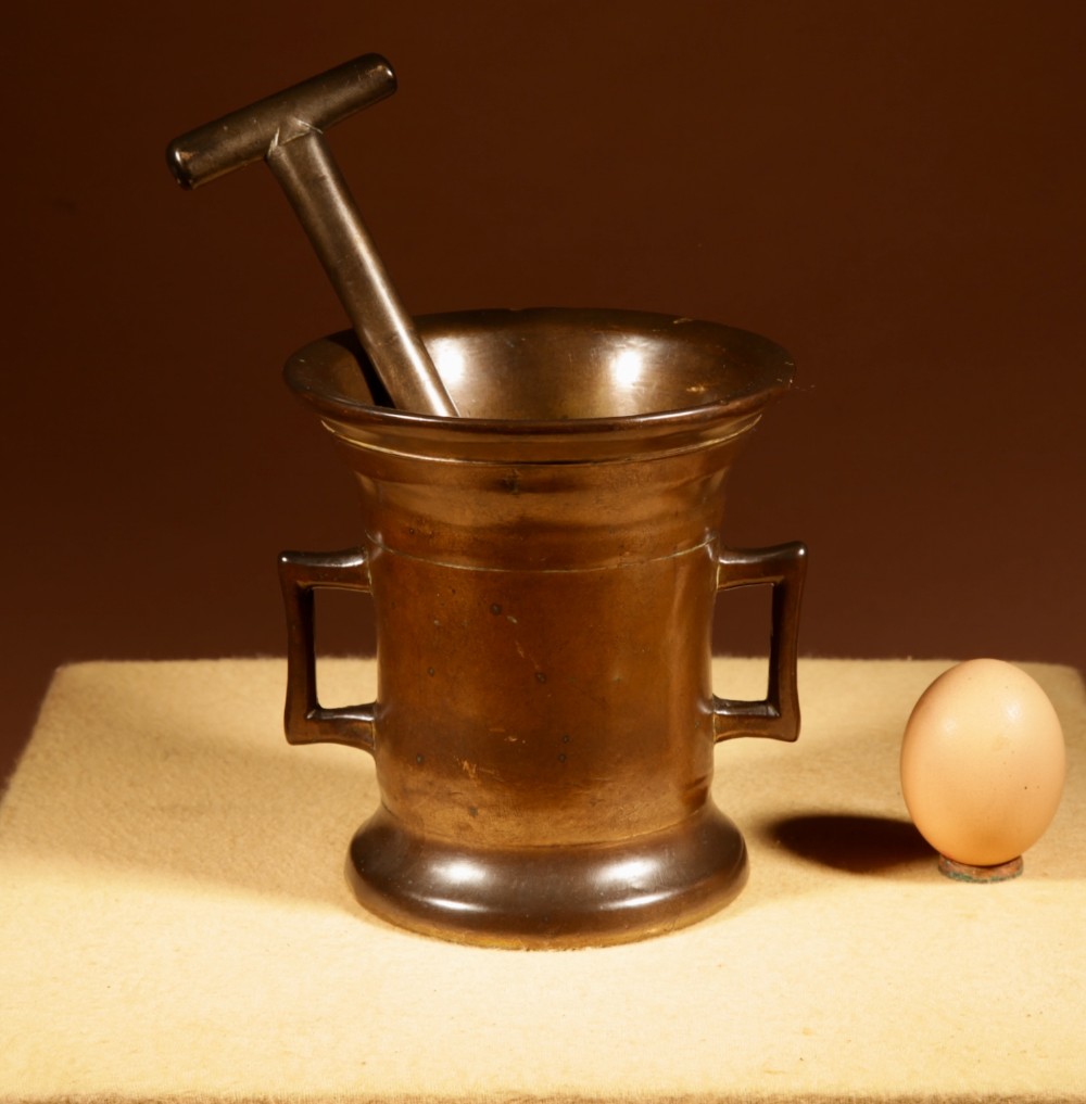A heavy cast bronze mortar featuring twin side handles and a flared rim, shown alongside its original T-bar pestle.