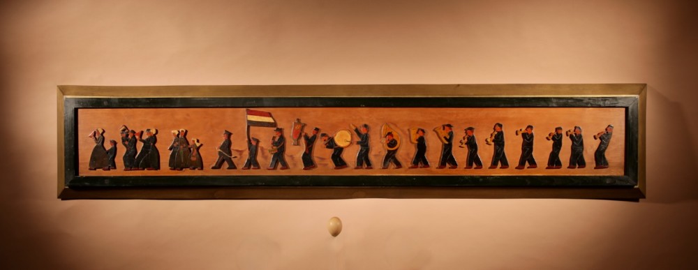 A long horizontal framed wooden relief panel depicting a painted Dutch marching band procession.