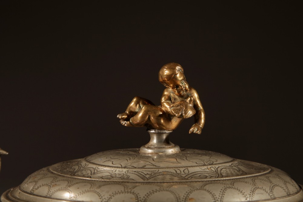 An 18th-century pewter birthing bowl featuring engraved floral patterns, twin scroll handles, ball feet, and a brass cherub finial on the lid. View II