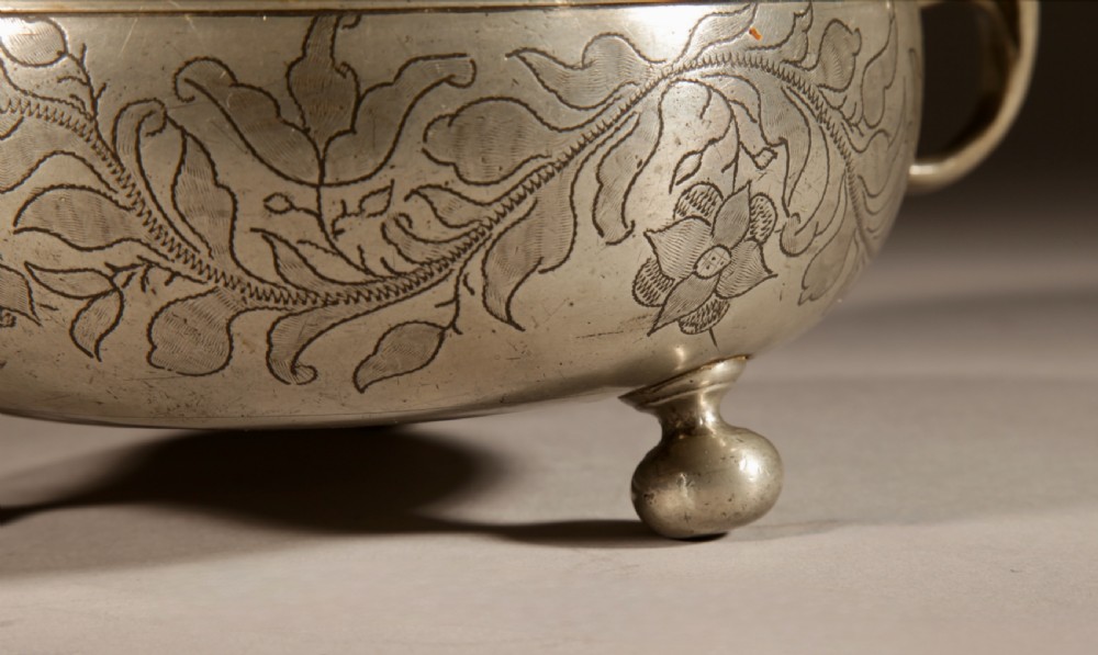 An 18th-century pewter birthing bowl featuring engraved floral patterns, twin scroll handles, ball feet, and a brass cherub finial on the lid. View III