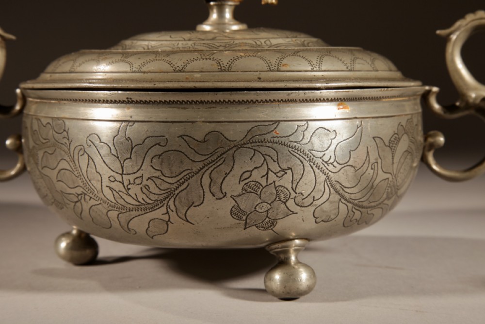An 18th-century pewter birthing bowl featuring engraved floral patterns, twin scroll handles, ball feet, and a brass cherub finial on the lid. View IV