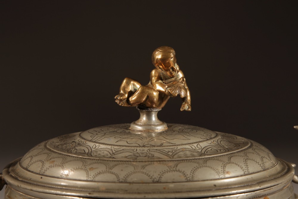 An 18th-century pewter birthing bowl featuring engraved floral patterns, twin scroll handles, ball feet, and a brass cherub finial on the lid. View V