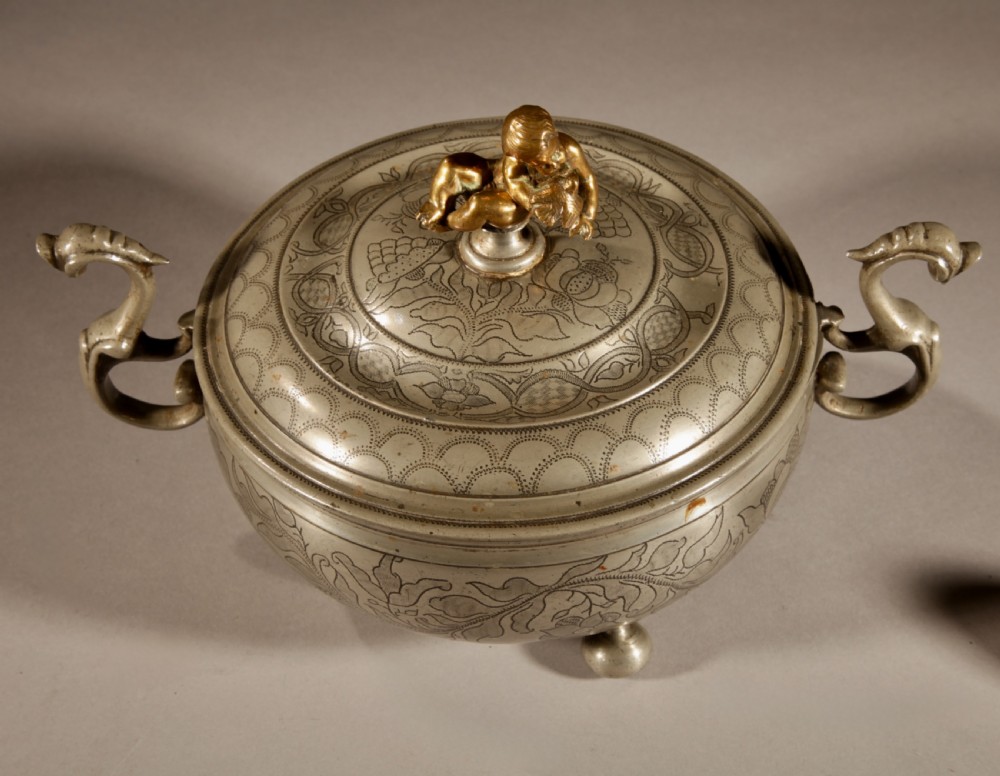 An 18th-century pewter birthing bowl featuring engraved floral patterns, twin scroll handles, ball feet, and a brass cherub finial on the lid. View VI
