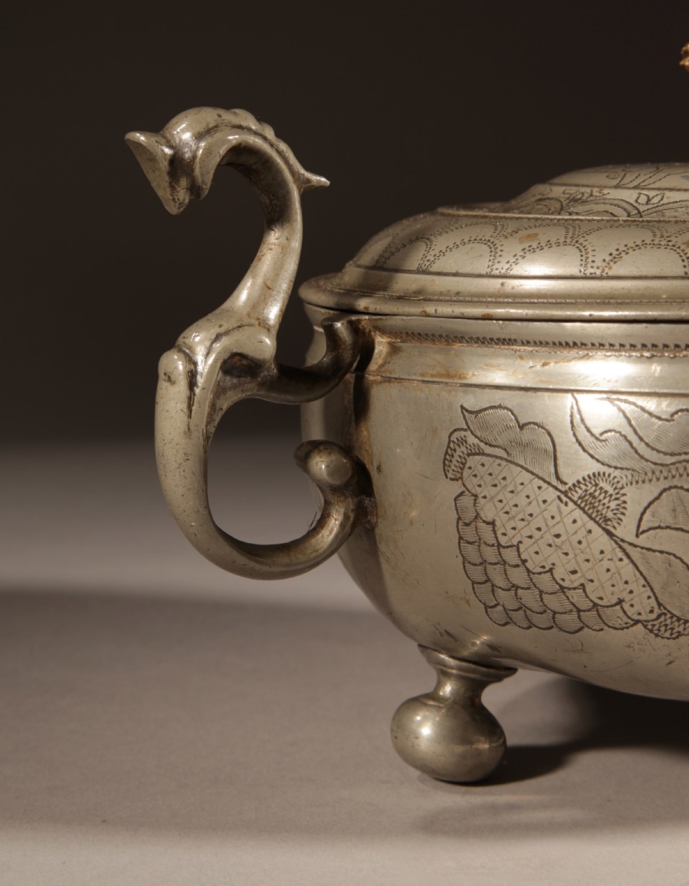 An 18th-century pewter birthing bowl featuring engraved floral patterns, twin scroll handles, ball feet, and a brass cherub finial on the lid. View VII