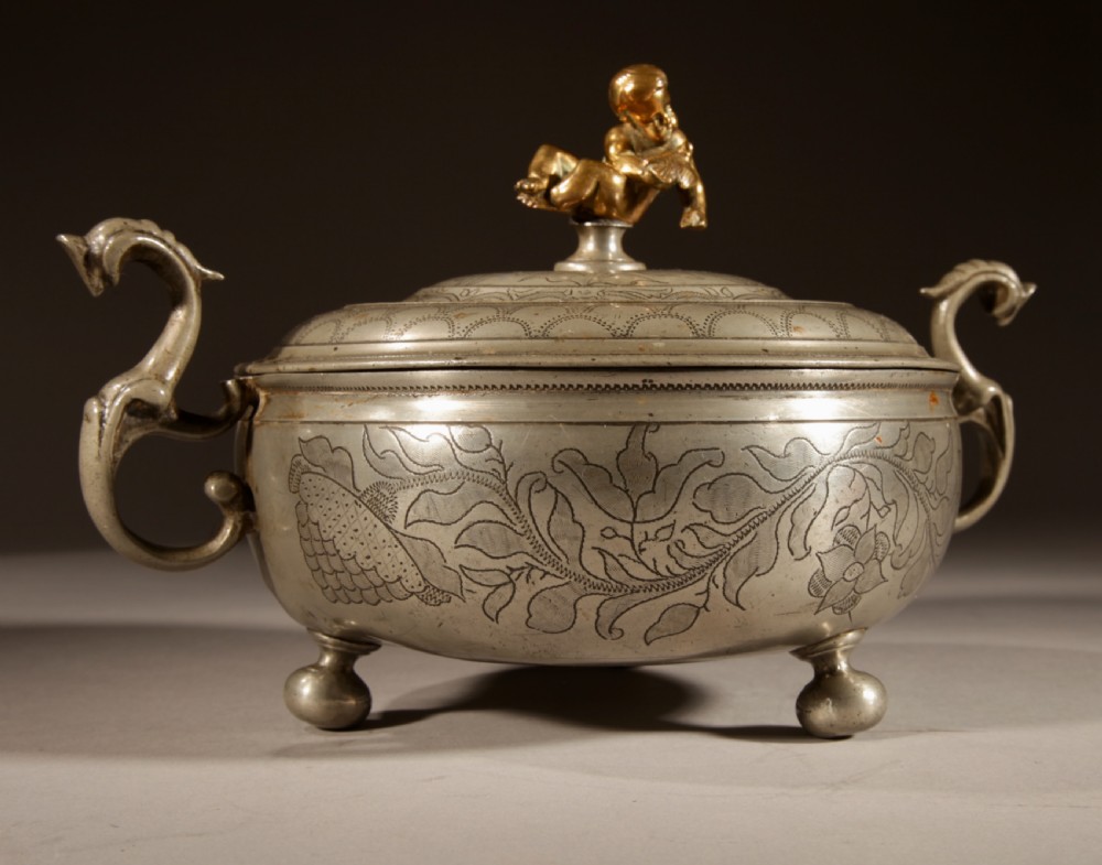 An 18th-century pewter birthing bowl featuring engraved floral patterns, twin scroll handles, ball feet, and a brass cherub finial on the lid. View VIII