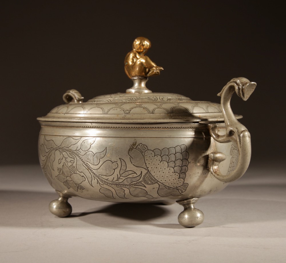 An 18th-century pewter birthing bowl featuring engraved floral patterns, twin scroll handles, ball feet, and a brass cherub finial on the lid. View IX