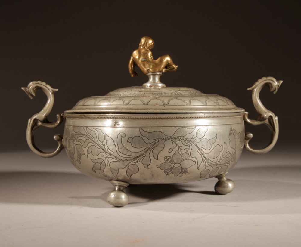 An 18th-century pewter birthing bowl featuring engraved floral patterns, twin scroll handles, ball feet, and a brass cherub finial on the lid. View X