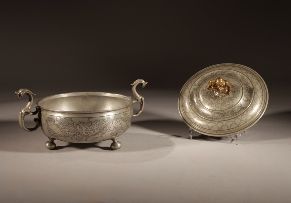 An 18th-century pewter birthing bowl featuring engraved floral patterns, twin scroll handles, ball feet, and a brass cherub finial on the lid. View XI