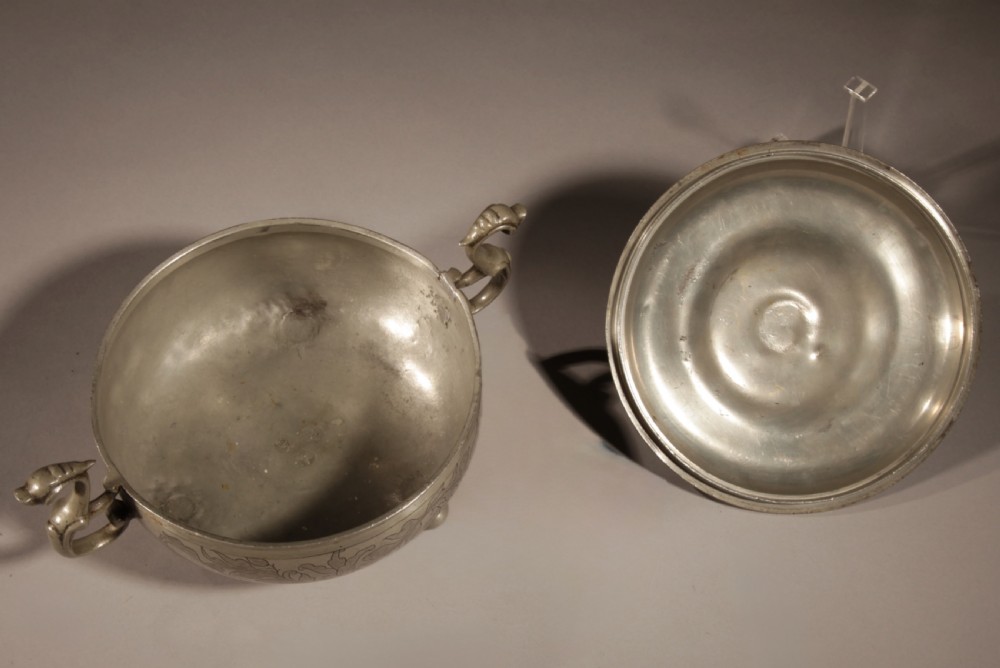 An 18th-century pewter birthing bowl featuring engraved floral patterns, twin scroll handles, ball feet, and a brass cherub finial on the lid. View XIII