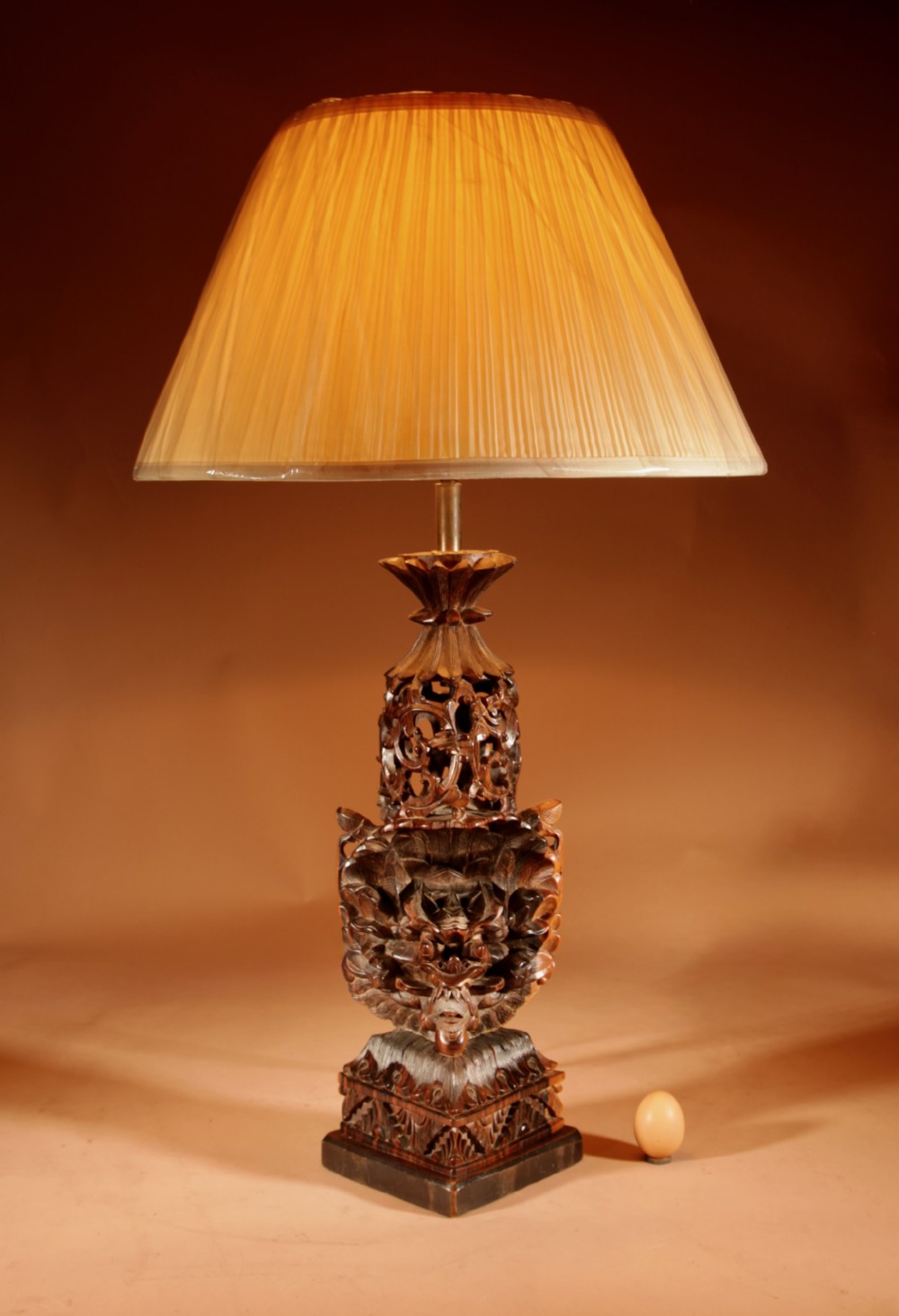 A carved dark hardwood table lamp featuring a detailed mythological mask motif and pierced lattice section, topped with a pleated beige fabric shade.