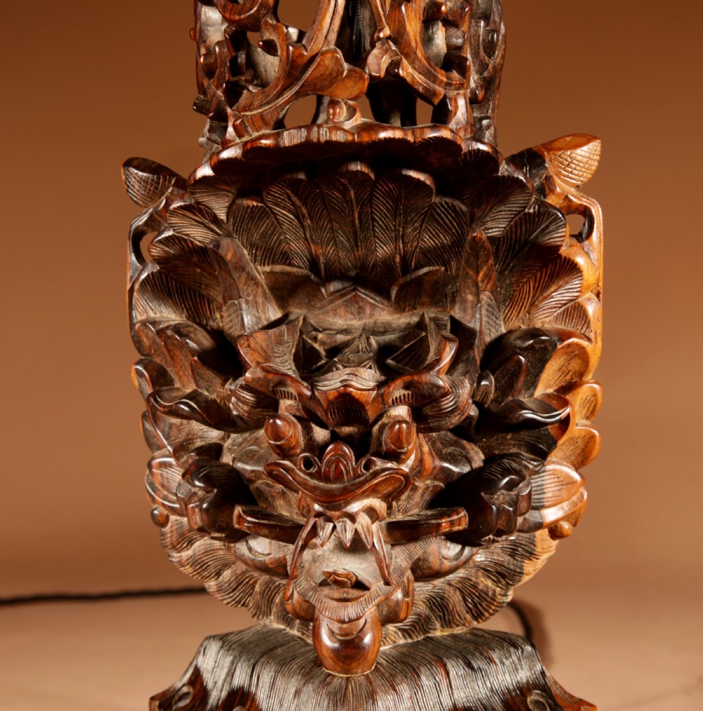 A carved dark hardwood table lamp featuring a detailed mythological mask motif and pierced lattice section, topped with a pleated beige fabric shade. View IV