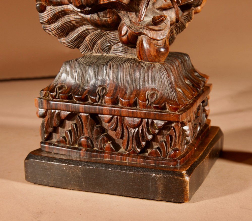 A carved dark hardwood table lamp featuring a detailed mythological mask motif and pierced lattice section, topped with a pleated beige fabric shade. View V