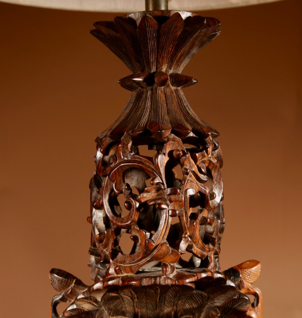 A carved dark hardwood table lamp featuring a detailed mythological mask motif and pierced lattice section, topped with a pleated beige fabric shade. View VII