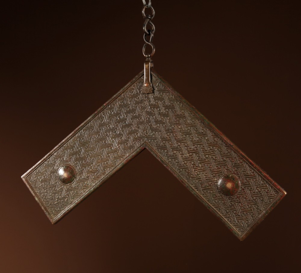 A chevron-shaped cast bronze musical chime featuring geometric relief patterns and a top suspension loop. View VIII