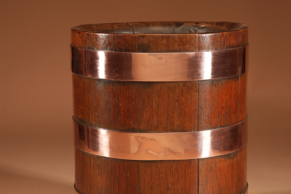 A cylindrical dark oak planter constructed with vertical timber staves, bound by three horizontal copper bands, and supported by small wooden feet. View VI
