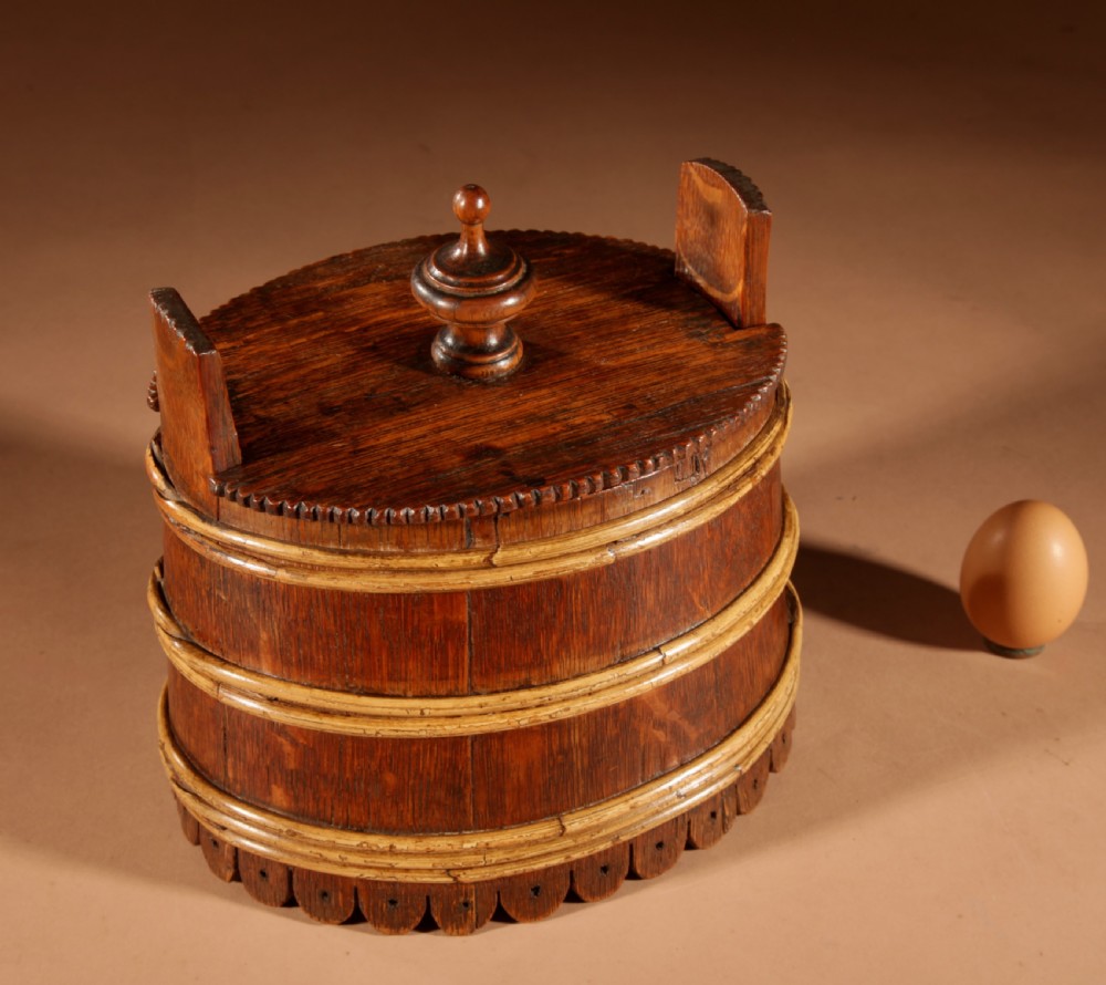 An oval oak butter tub featuring coopered staves, bound wood hoops, two raised side handles, a carved scalloped base, and a fitted lid with a turned central finial. View III