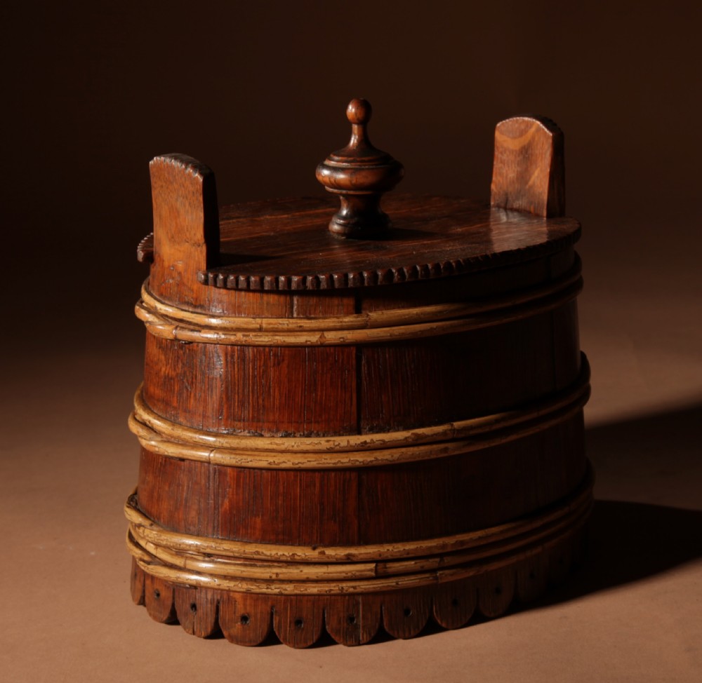 An oval oak butter tub featuring coopered staves, bound wood hoops, two raised side handles, a carved scalloped base, and a fitted lid with a turned central finial. View IV