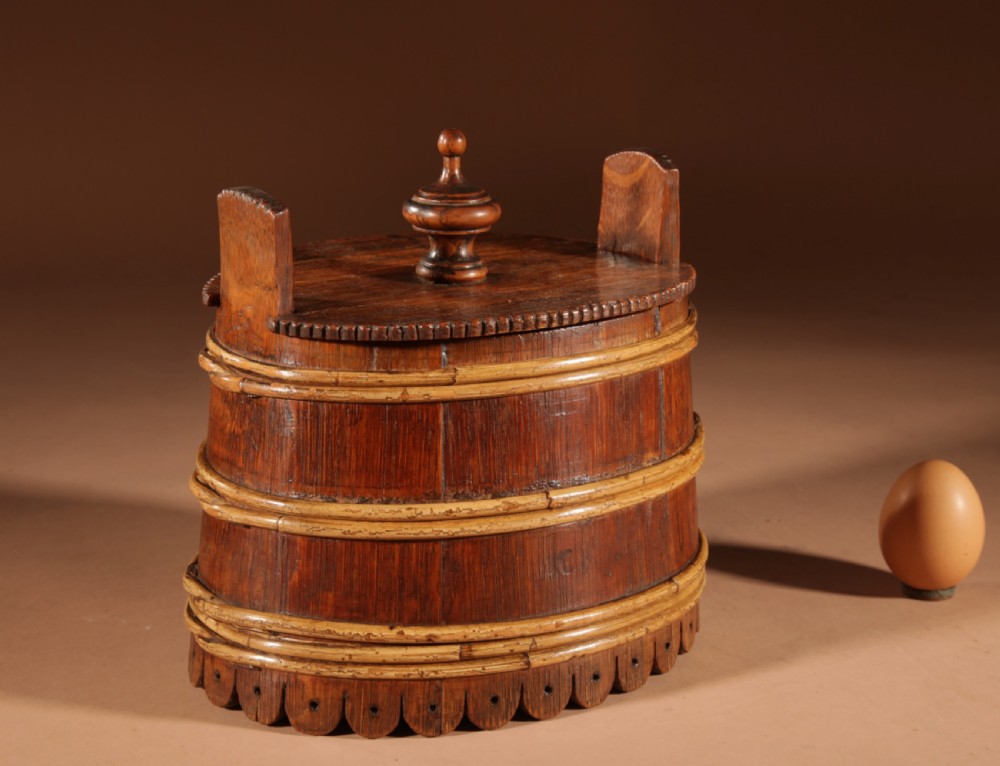 An oval oak butter tub featuring coopered staves, bound wood hoops, two raised side handles, a carved scalloped base, and a fitted lid with a turned central finial. View VI
