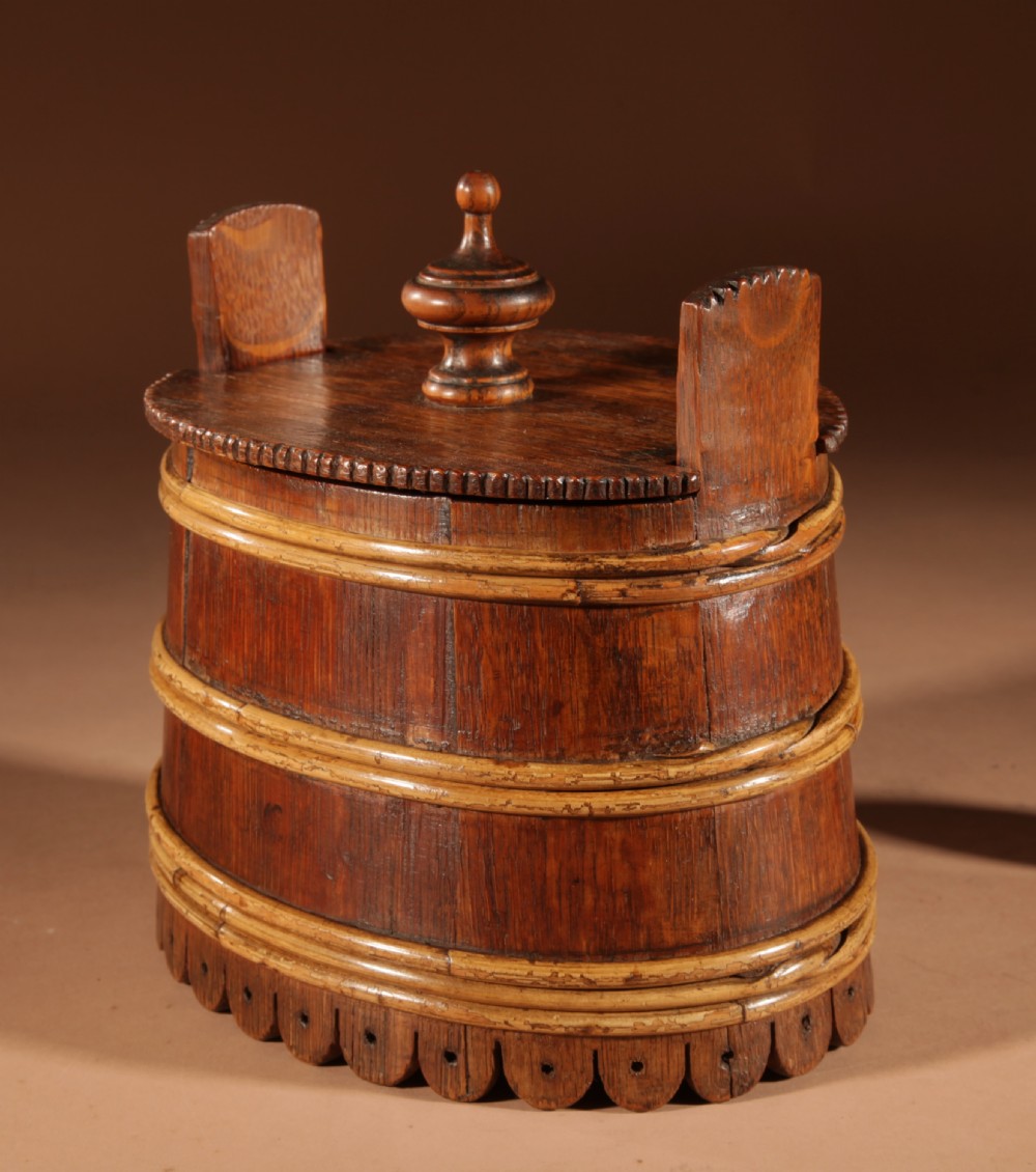 An oval oak butter tub featuring coopered staves, bound wood hoops, two raised side handles, a carved scalloped base, and a fitted lid with a turned central finial. View VII
