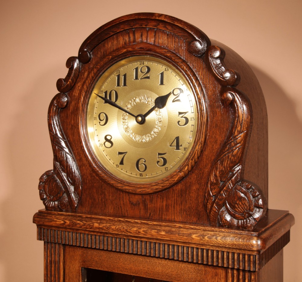 A dark oak longcase clock featuring carved arch cresting, a brass dial with Arabic numerals, and a glass trunk door displaying brass weights and pendulum. View IV